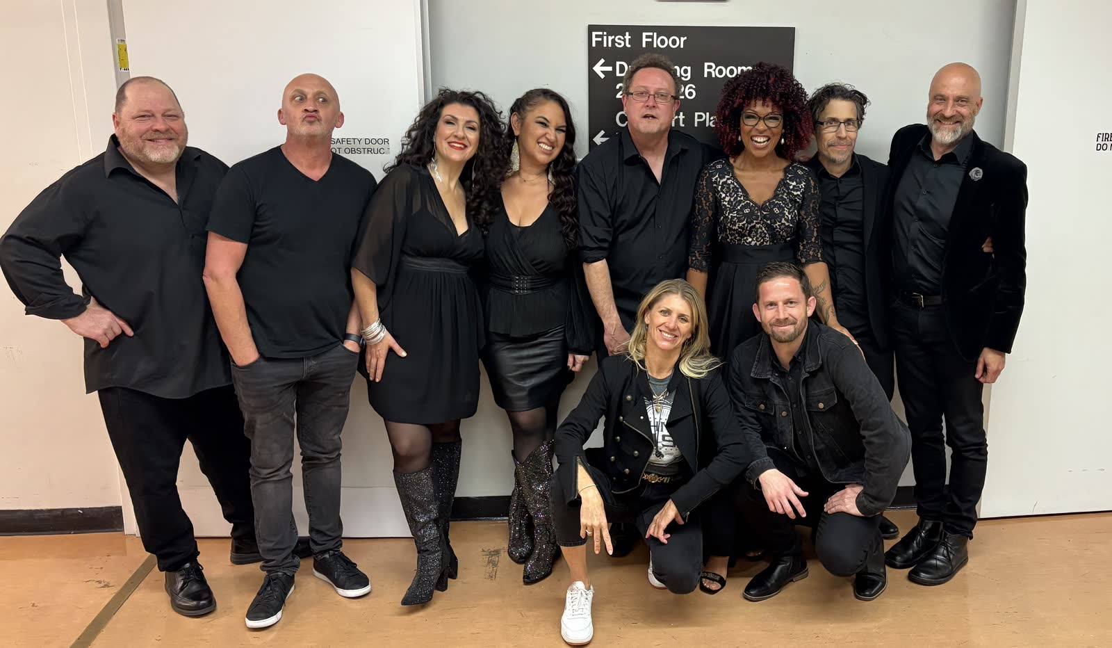 Ali Winton crouching at the front of a group of musicians and crew dressed in black, backstage in a venue corridor beneath a sign reading First Floor, Dressing Rooms