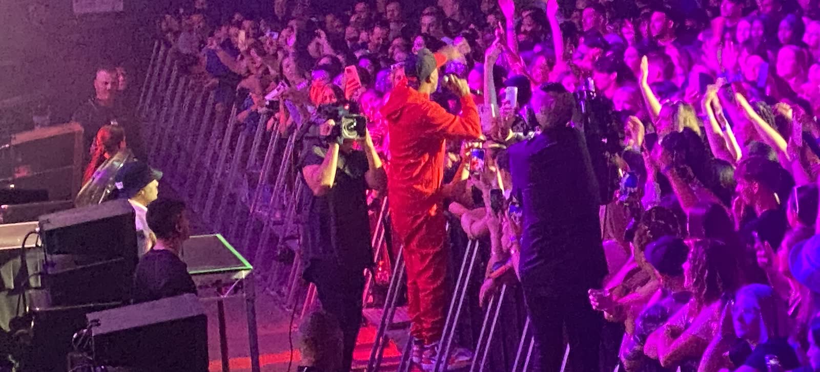 Front of house at a packed show: a performer in a red boiler suit leaning over the barrier into the crowd, with a camera operator, security and rows of raised phones